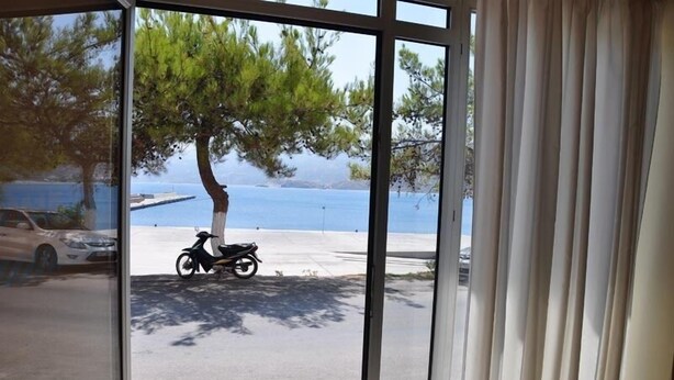 A view through a window of a coastal area with a scooter parked on the street, a tree, and a body of water in the background. - Nora Hotel (Sitia)