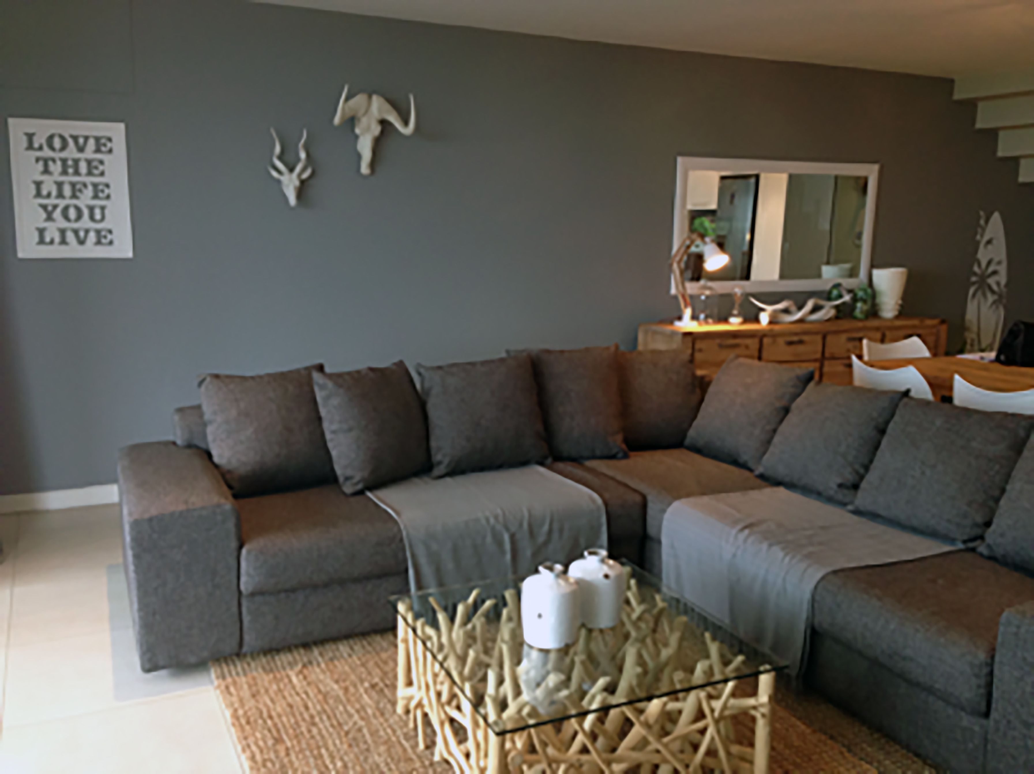 A modern living room with a grey sectional sofa, a glass coffee table with decorative items, a mirror, and a wall-mounted sign.