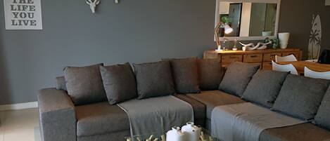 A modern living room with a grey sectional sofa, a glass coffee table with decorative items, a mirror, and a wall-mounted sign.