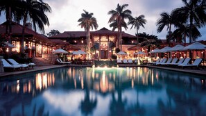A pool area with lounge chairs and umbrellas, surrounded by palm trees and a building with a red roof. - Zimbali Condo (Ballito)