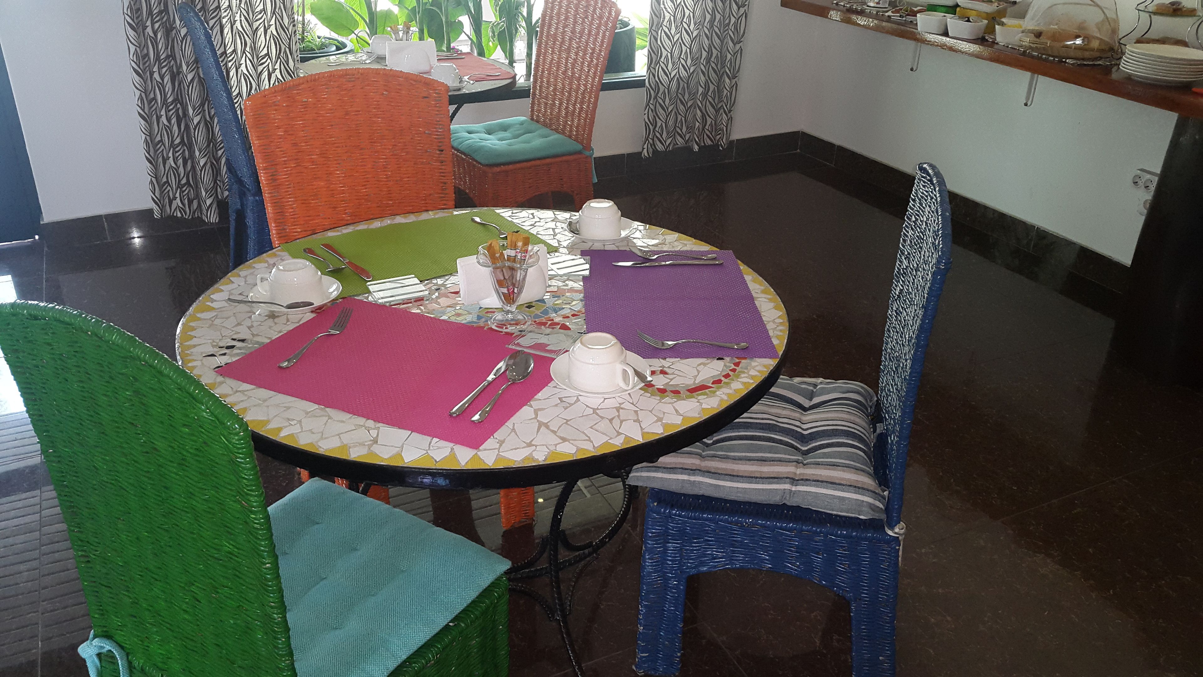Un comedor con una mesa redonda preparada para dos comensales, con mantel de mosaico, sillas de colores y una variedad de vajilla.