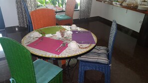 A dining area with a round table set for two, featuring a mosaic tablecloth, colorful chairs, and a variety of tableware. - Mozaika Guesthouse 2 (Maputo)