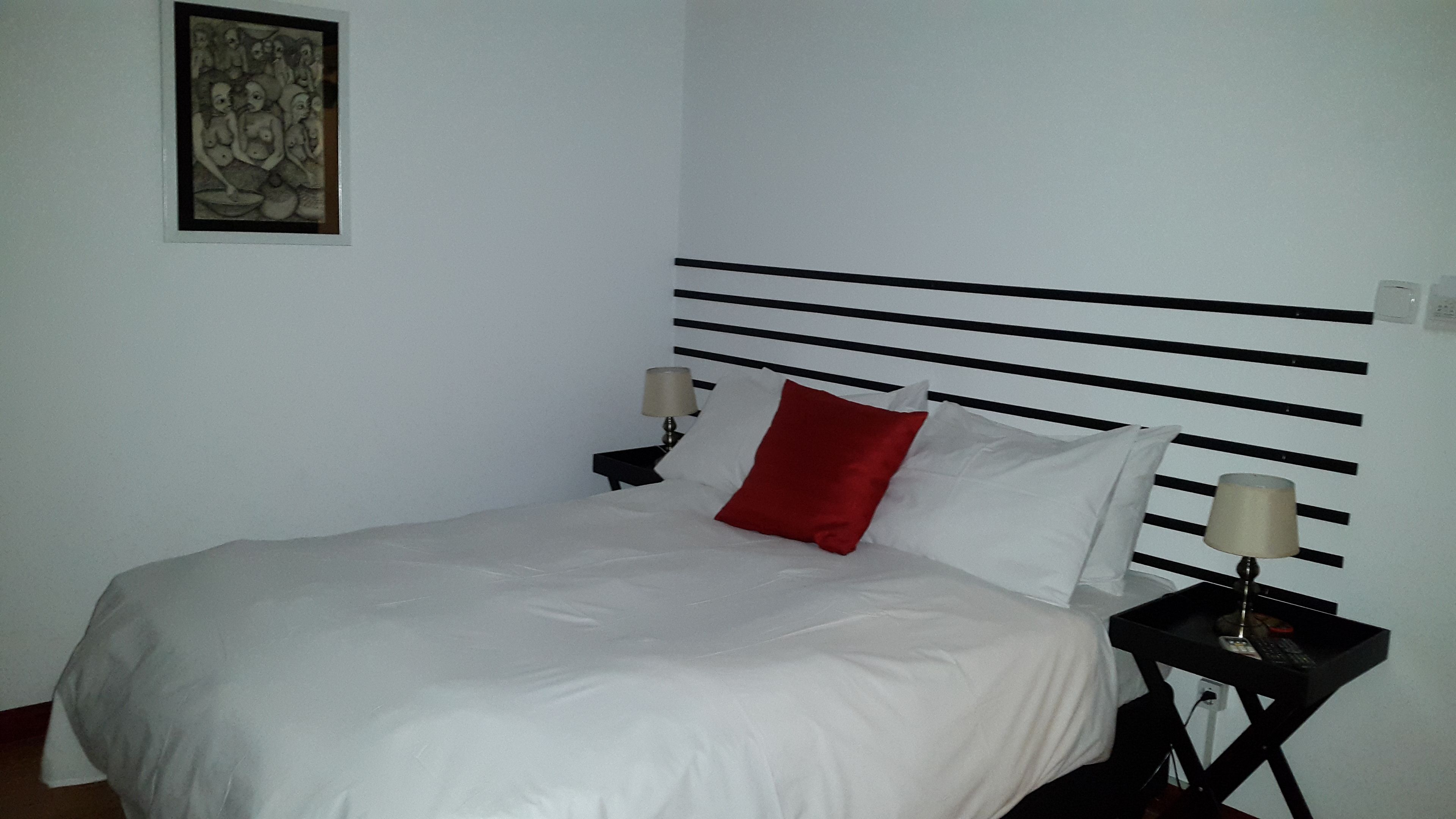 A neatly made bed with white bedding and a red pillow, a black headboard with horizontal lines, two bedside tables with lamps, and a framed artwork on the wall.