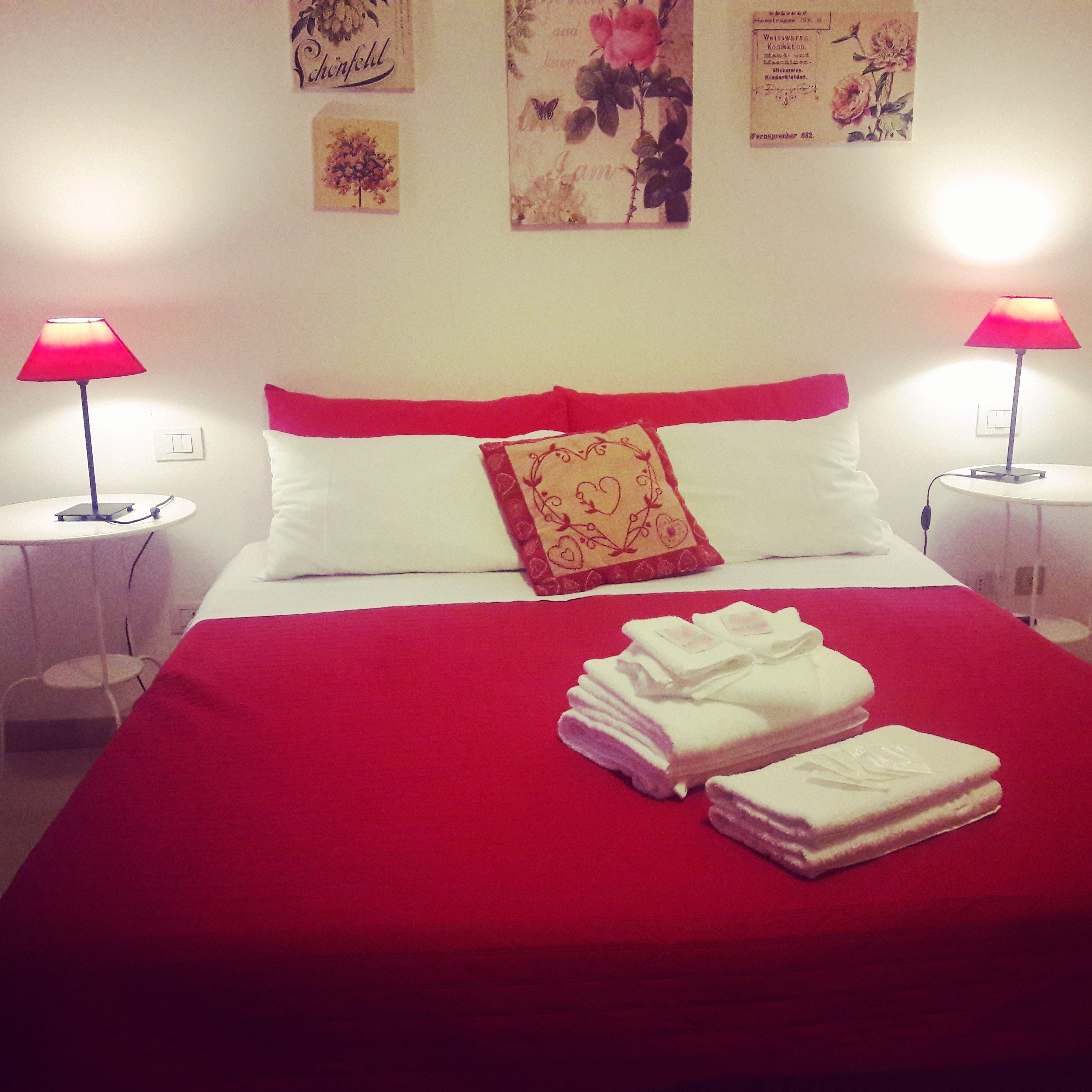 Una cama bien hecha con ropa de cama roja y almohadas blancas, dos lámparas de noche y una pared con obras de arte enmarcadas.