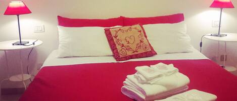 Una cama bien hecha con ropa de cama roja y almohadas blancas, dos lámparas de noche y una pared con obras de arte enmarcadas.