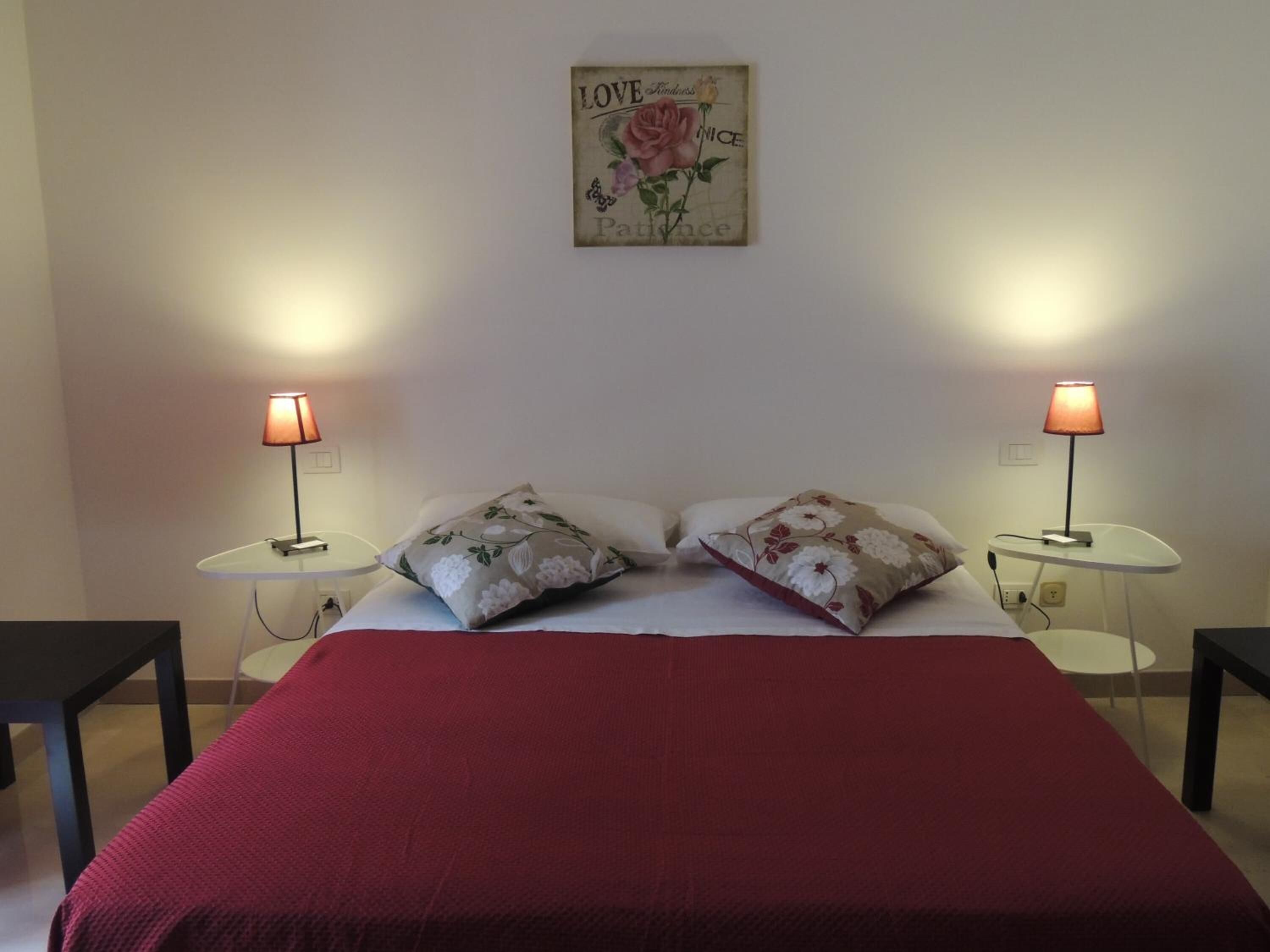 A neatly made bed with a red blanket, two floral pillows, and two bedside lamps on white nightstands.