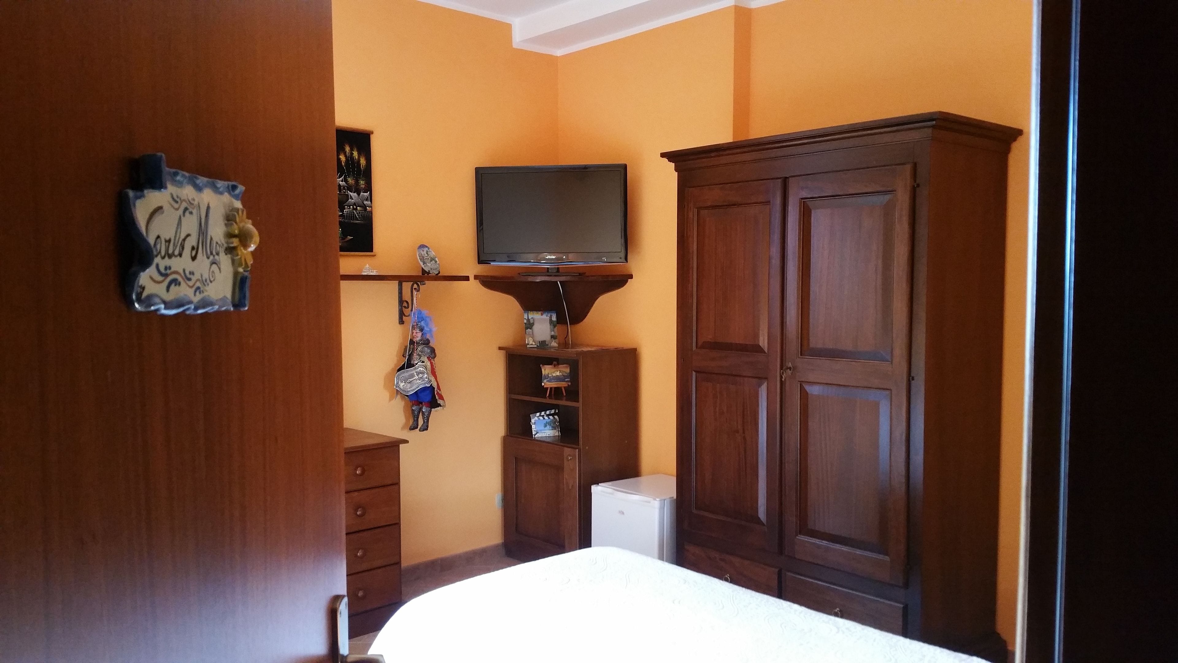 A bedroom with a bed, wooden wardrobe, a television on a shelf, and a small refrigerator.