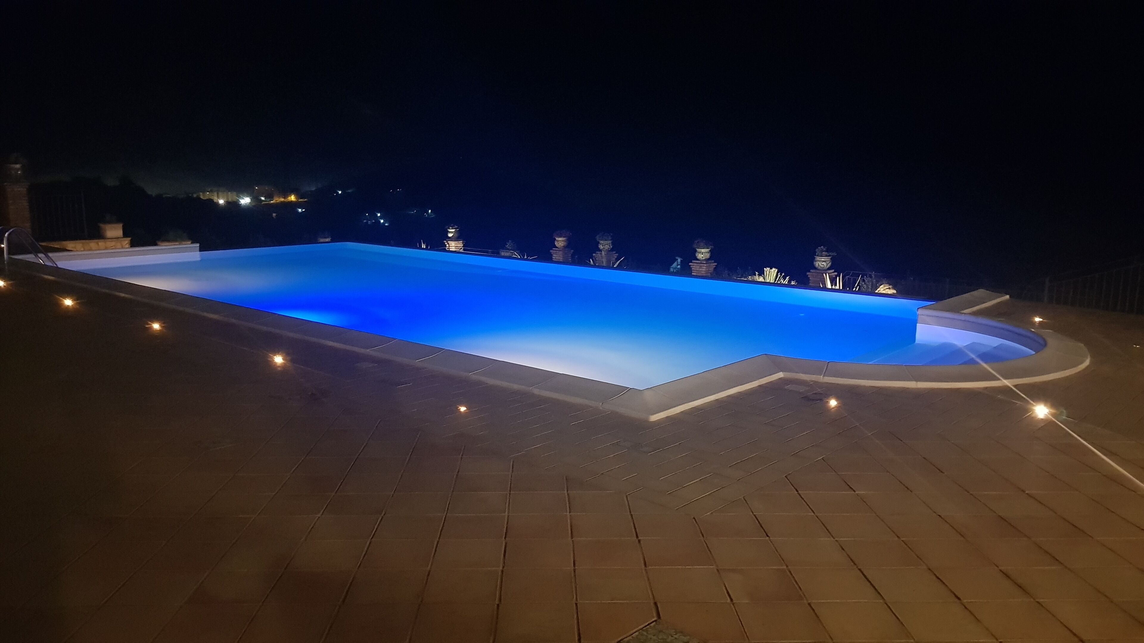 A rectangular swimming pool with a blue-lit edge, surrounded by a tiled deck with evenly spaced lights.