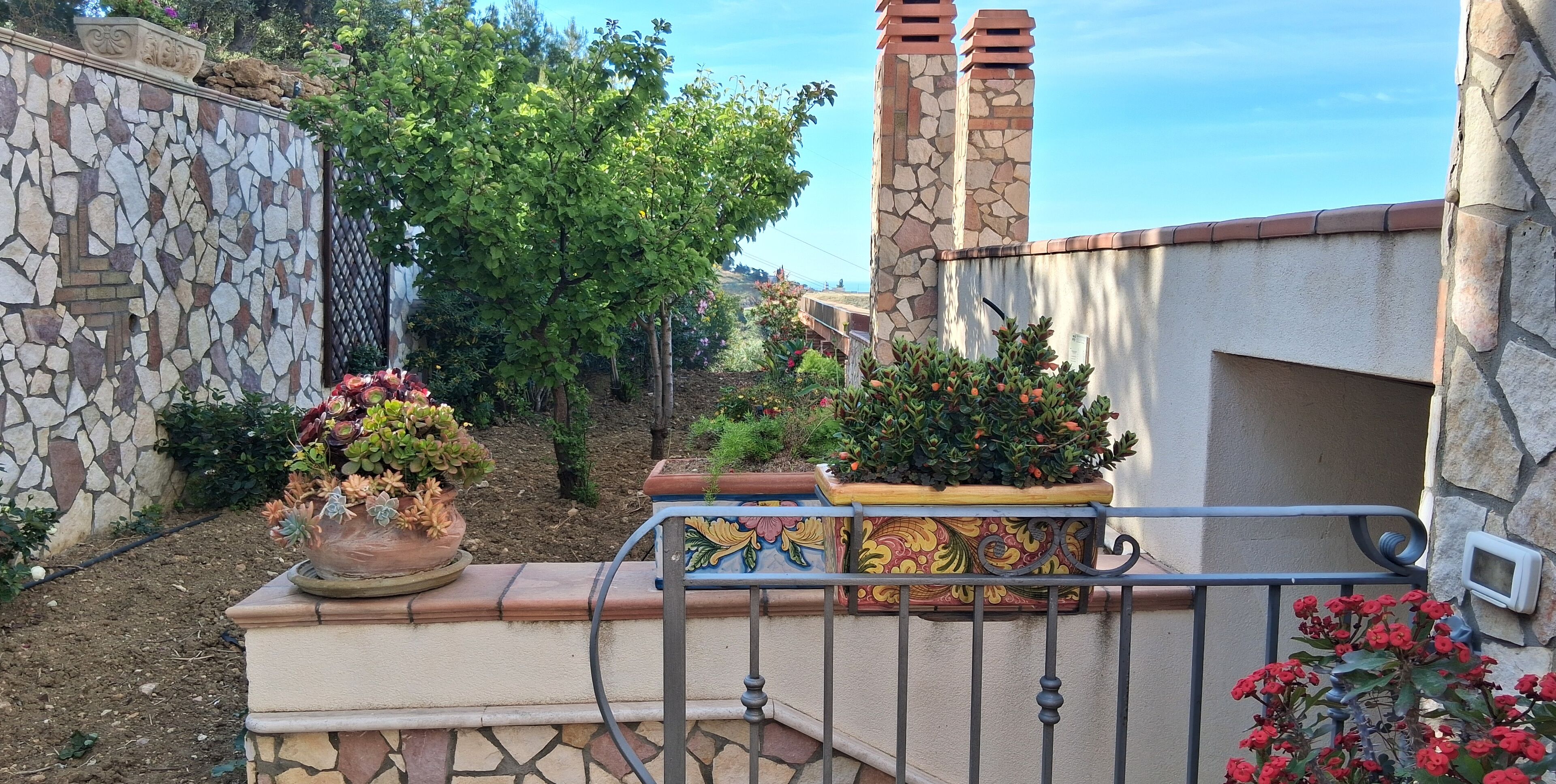 A stone wall with a decorative pattern, a potted plant with red flowers, and a metal railing.