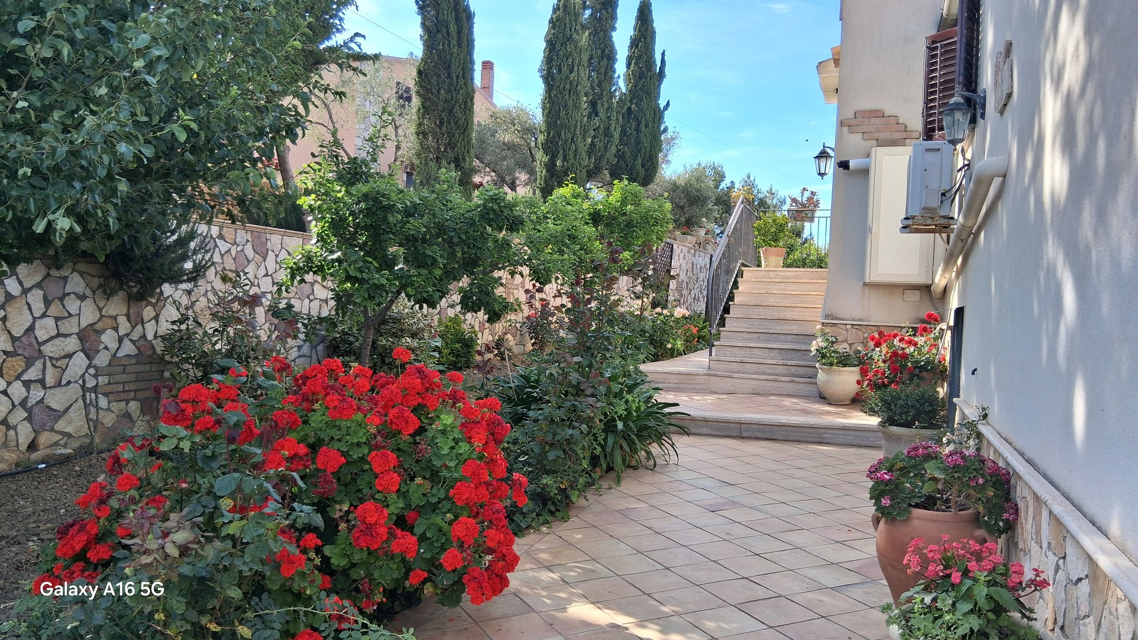 A garden pathway with red flowers, stone walls, and a staircase leading to a building.