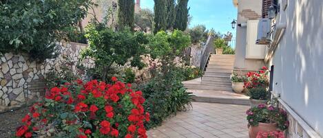 A garden pathway with red flowers, stone walls, and a staircase leading to a building.