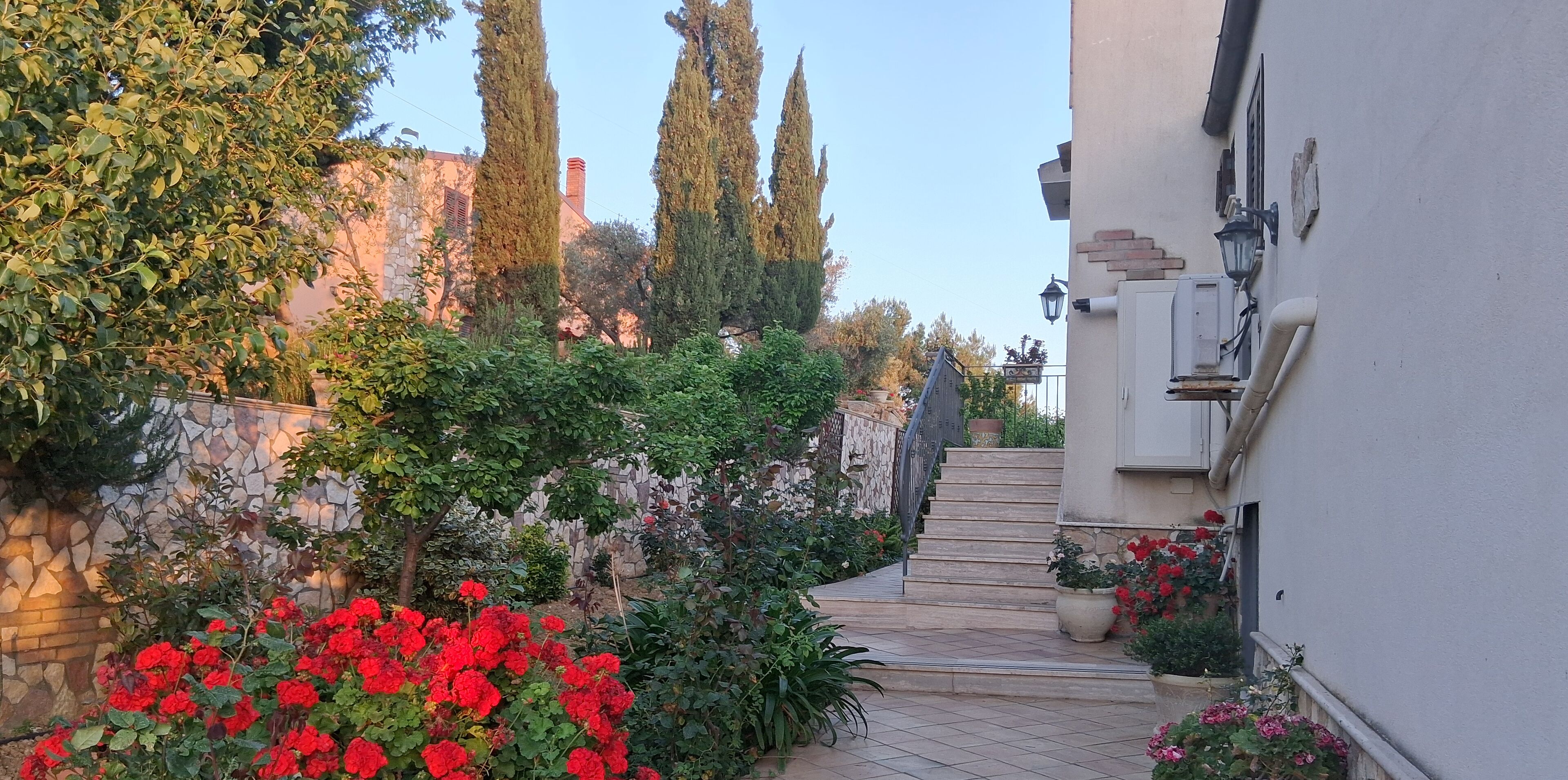 Un étroit sentier bordé d’une végétation luxuriante et de fleurs rouges mène à un bâtiment doté d’un mur en pierre et d’un escalier.