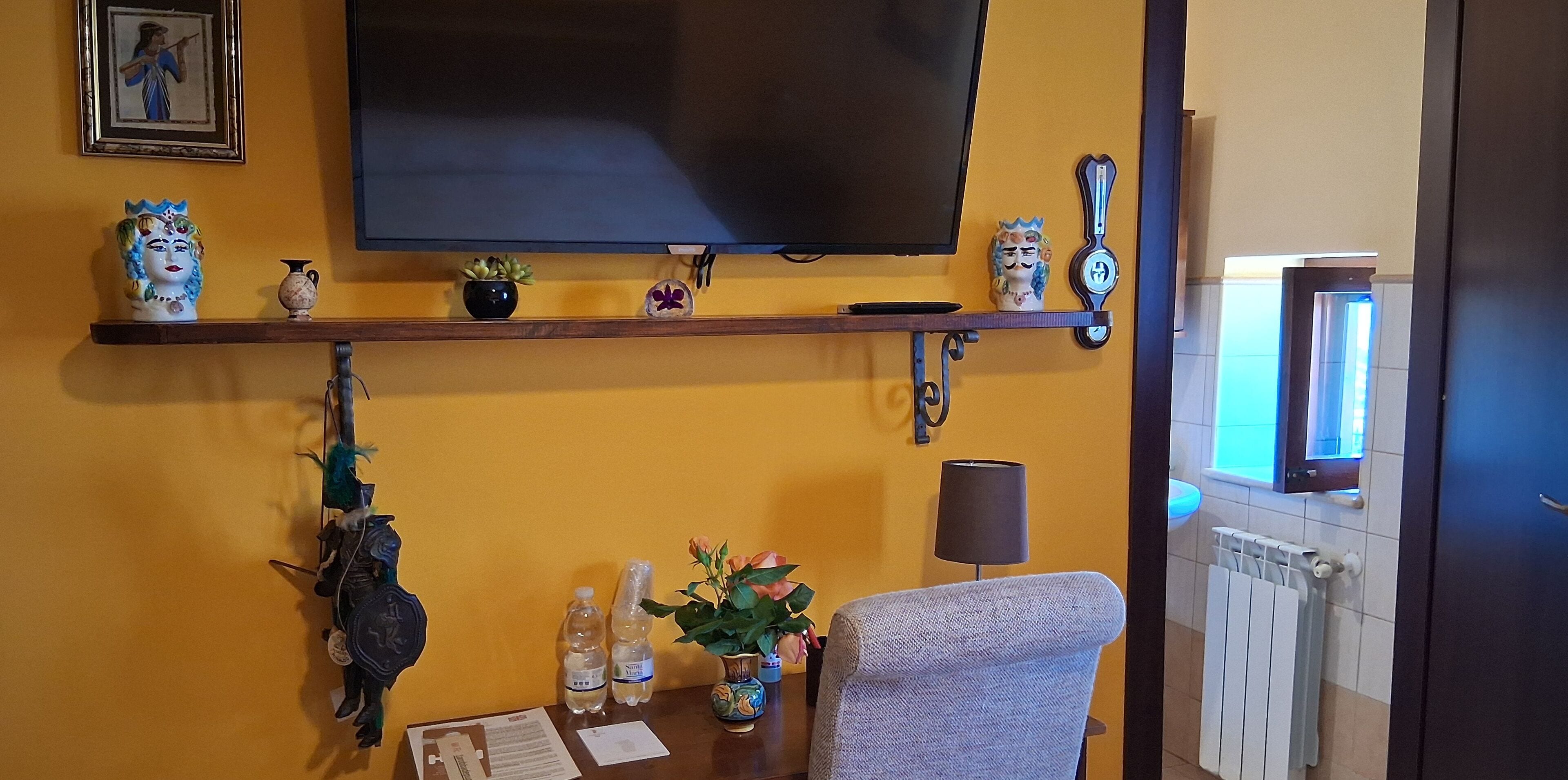 A living room with a mounted TV, decorative items on shelves, a table with papers, a chair, and a radiator.
