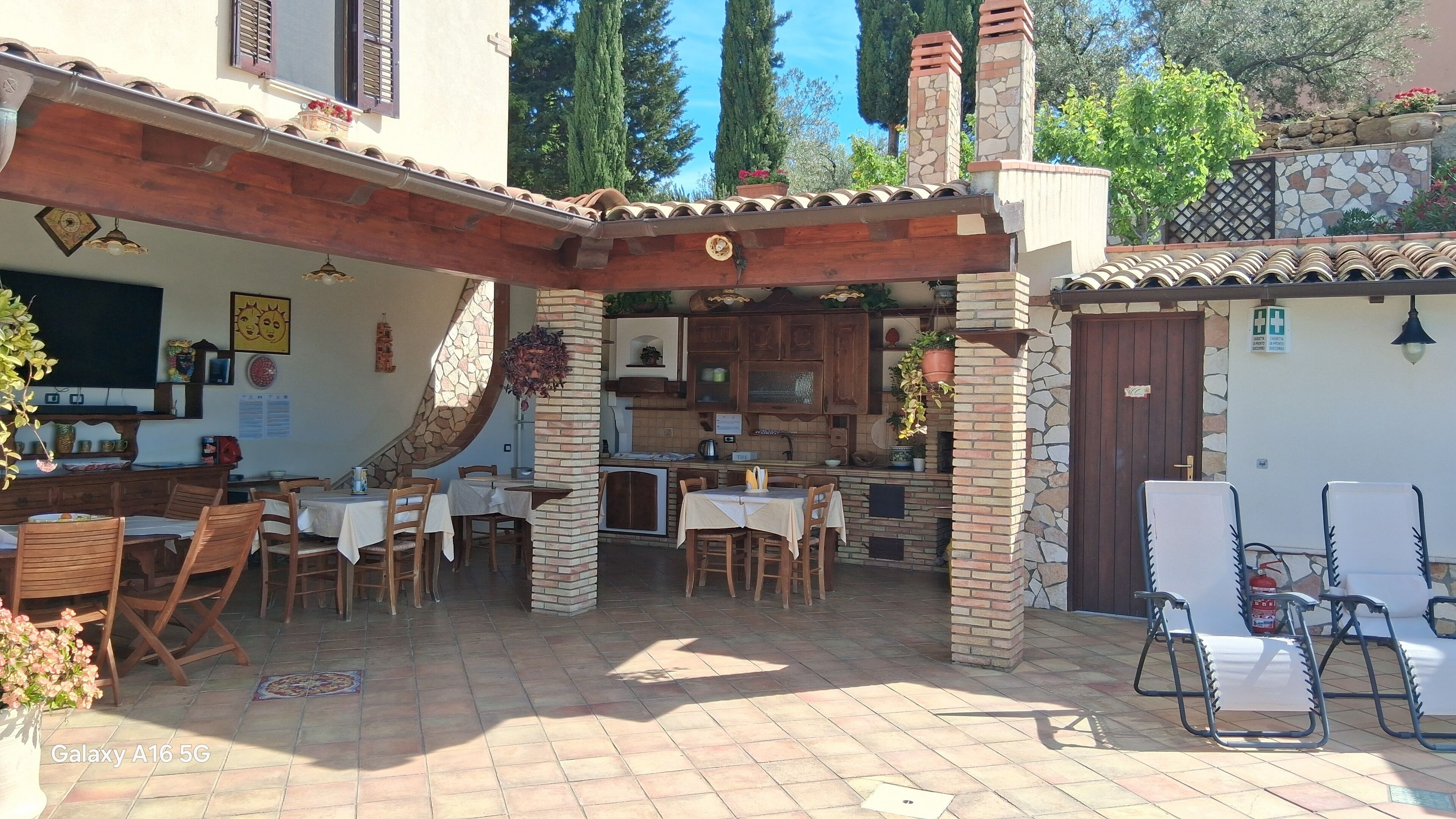 A patio area with wooden tables and chairs, a stone fireplace, and a kitchen area with wooden cabinets.