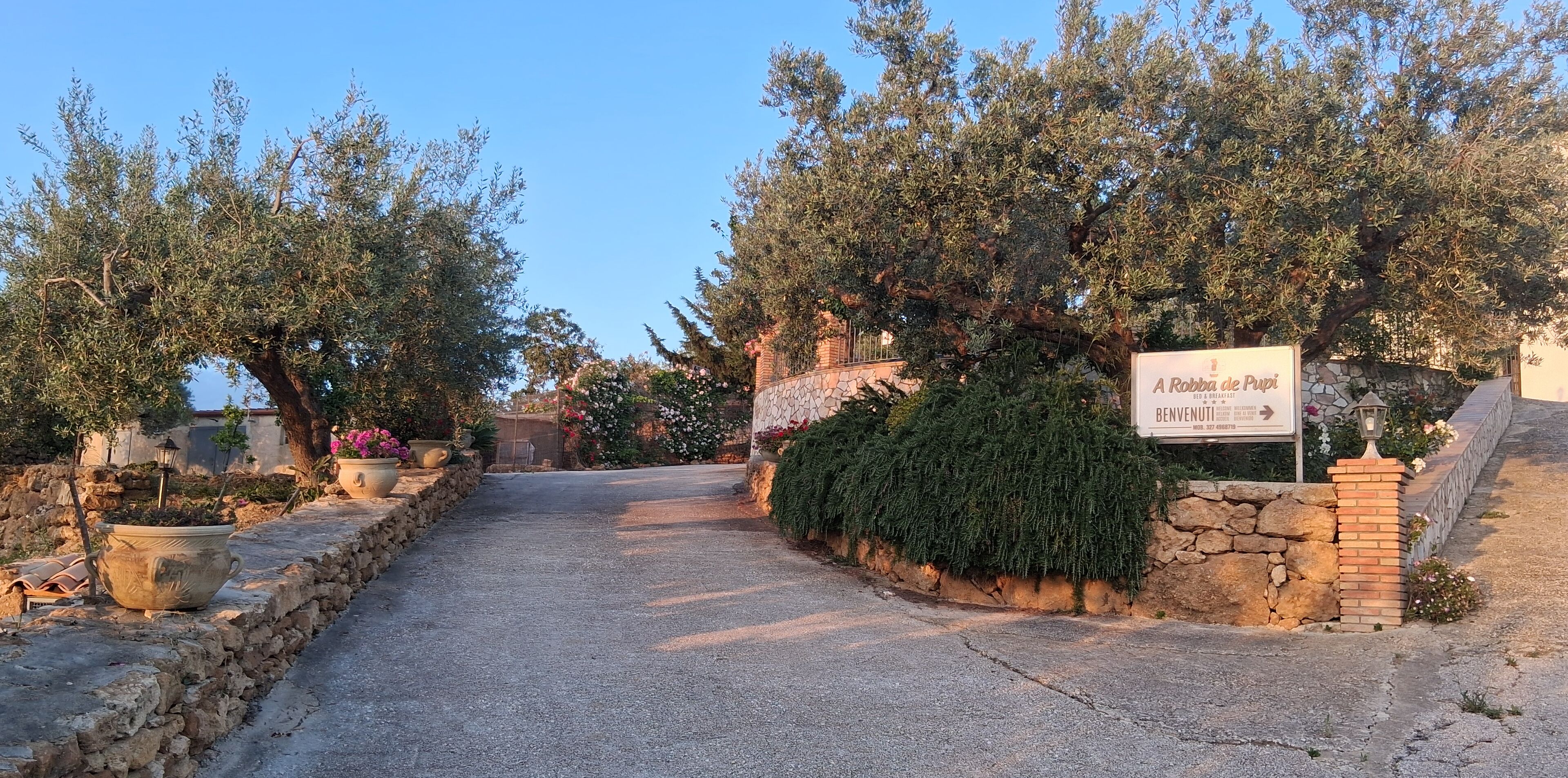 A stone pathway leading to a sign for 'Robba de Pupi' with a stone wall and potted plants on either side.