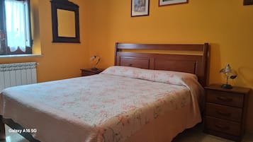 A bedroom with a bed, a nightstand, a dresser, a radiator, a window with curtains, and a wall-mounted mirror.