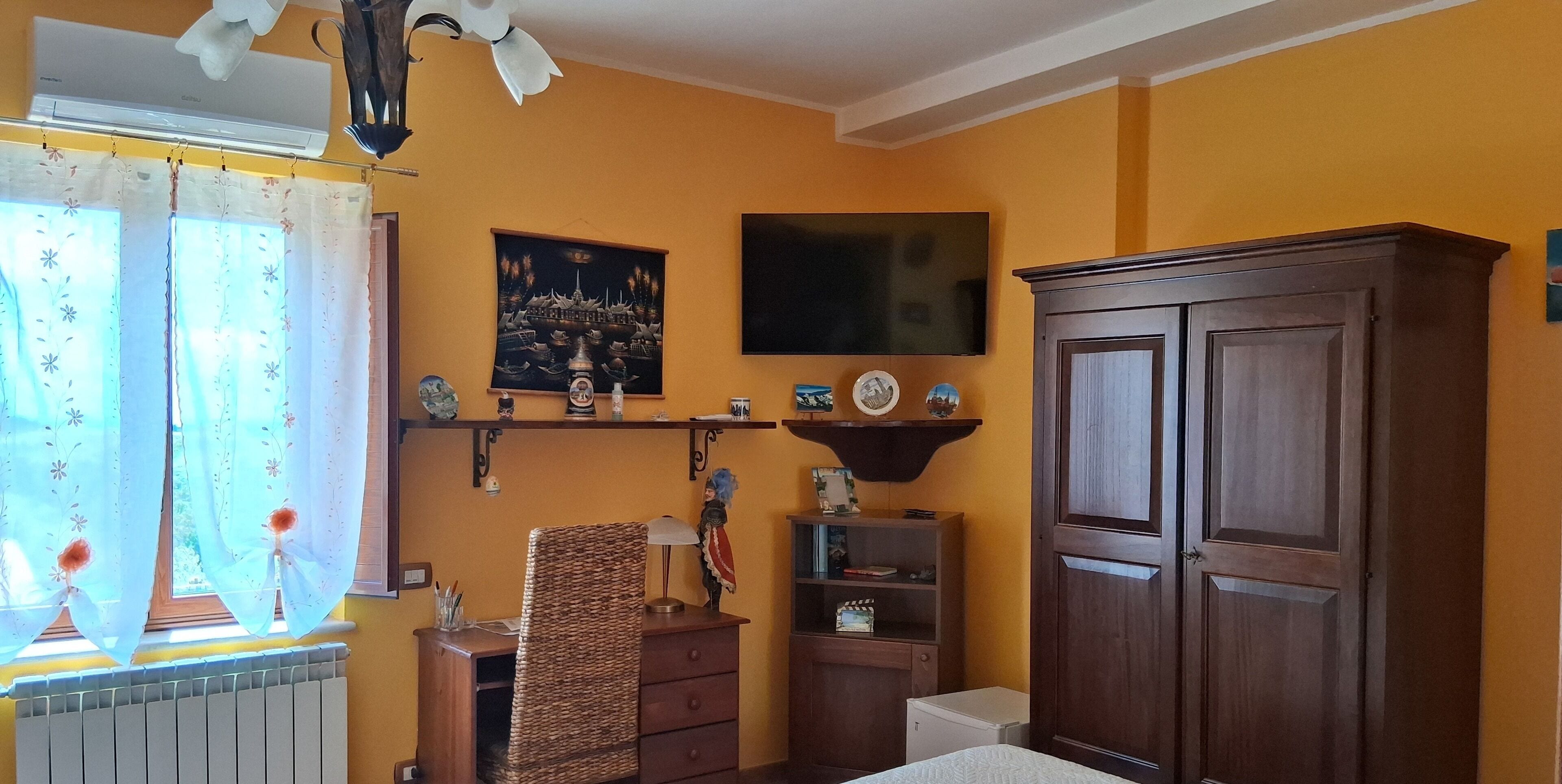 A room with a large wooden wardrobe, a small desk with a chair, a radiator, and a window with curtains.