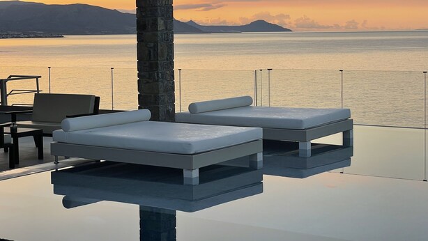 Ein moderner Außenbereich mit zwei weißen, gepolsterten Sofas und einem Tisch, mit Blick auf ein ruhiges Meer und ferne Berge im Abendrot.
