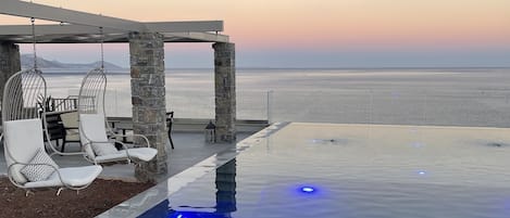 Una piscina infinita con vista al océano, pilares de piedra y sillones de descanso blancos.
