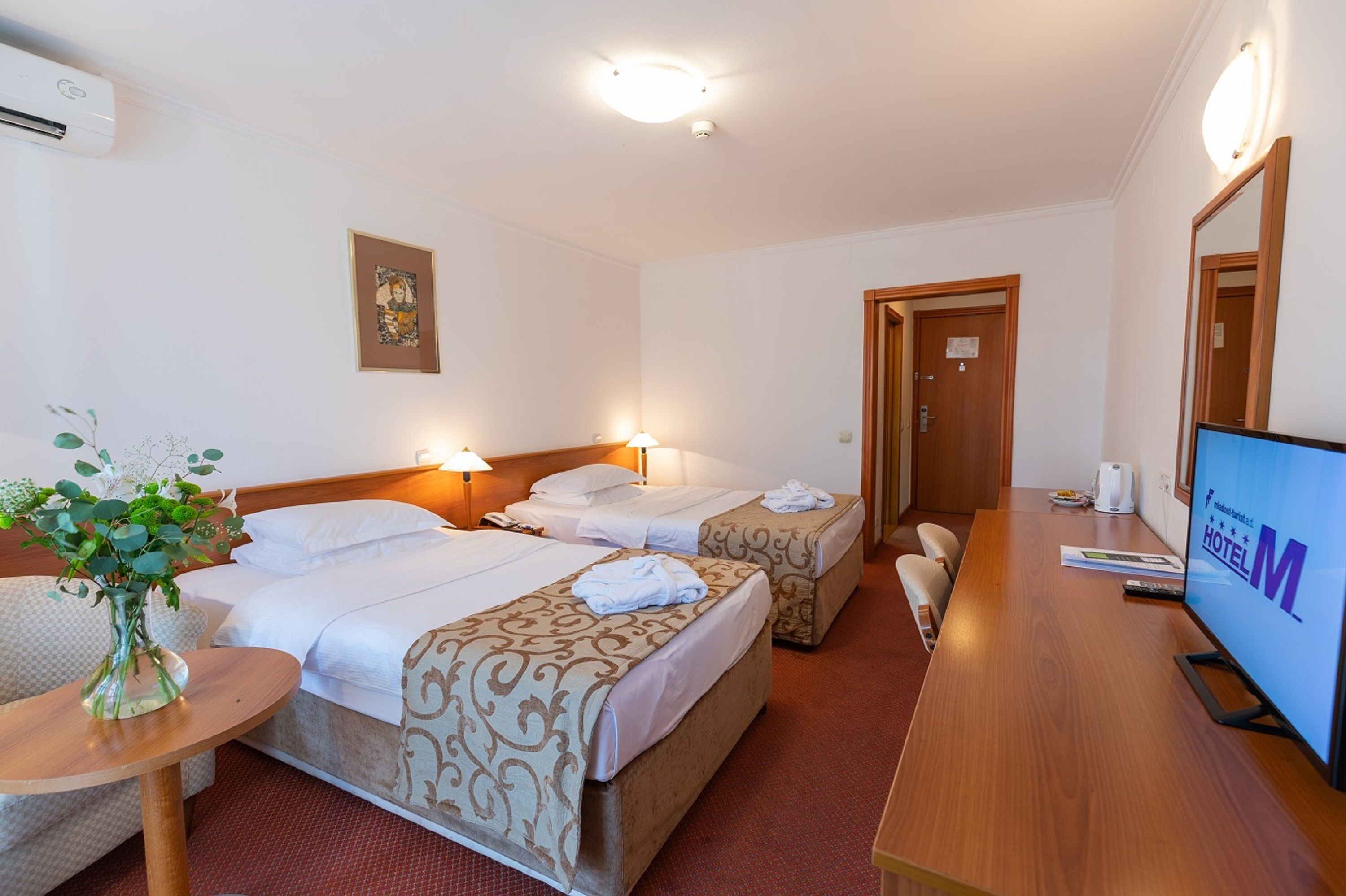 A hotel room with two beds, a TV, a desk, and a small table with a vase.