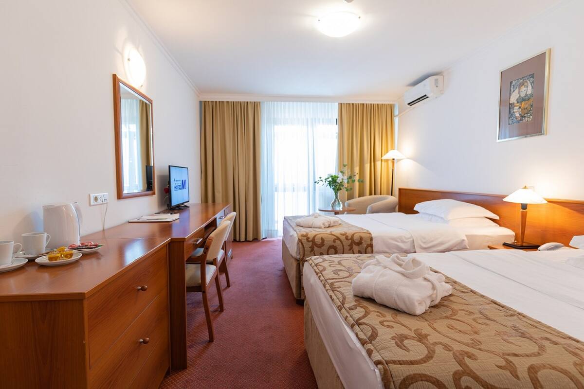 A hotel room with two beds, a desk with a computer, a chair, and a small table with a lamp.