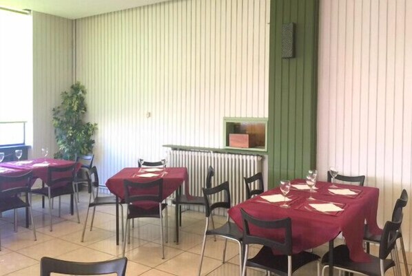 Un comedor con mesas dispuestas para cuatro, cada una con mantel rojo y sillas negras. Hay un radiador en la pared y una planta en maceta a la izquierda.