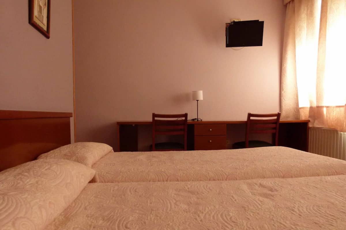A hotel room with two beds, a desk with two chairs, a lamp, and a small TV on the wall.