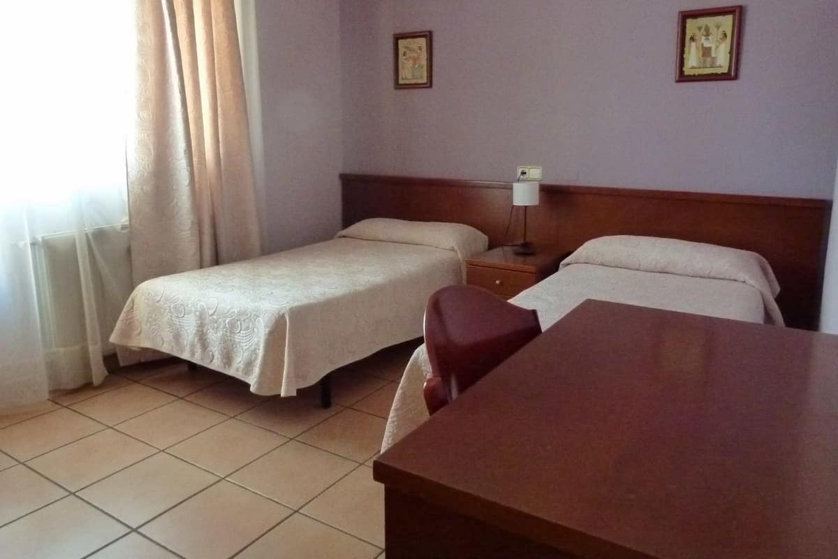 A hotel room with two single beds, a wooden desk, a nightstand with a lamp, and two framed pictures on the wall.