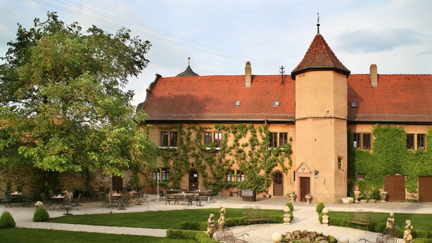 A building with a red-tiled roof, ivy-covered walls, and a central tower. There are outdoor tables and chairs, and a well-maintained garden with hedges and statues. - Wörners Schloss Weingut & Wellness Hotel (Prichsenstadt)