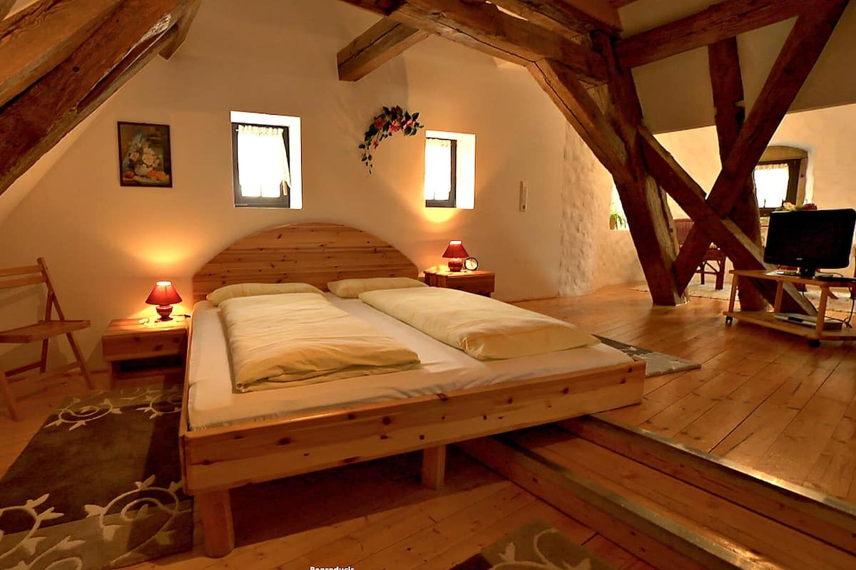 A cozy attic bedroom with a wooden bed, two bedside tables with lamps, a chair, and a TV on a wooden stand.