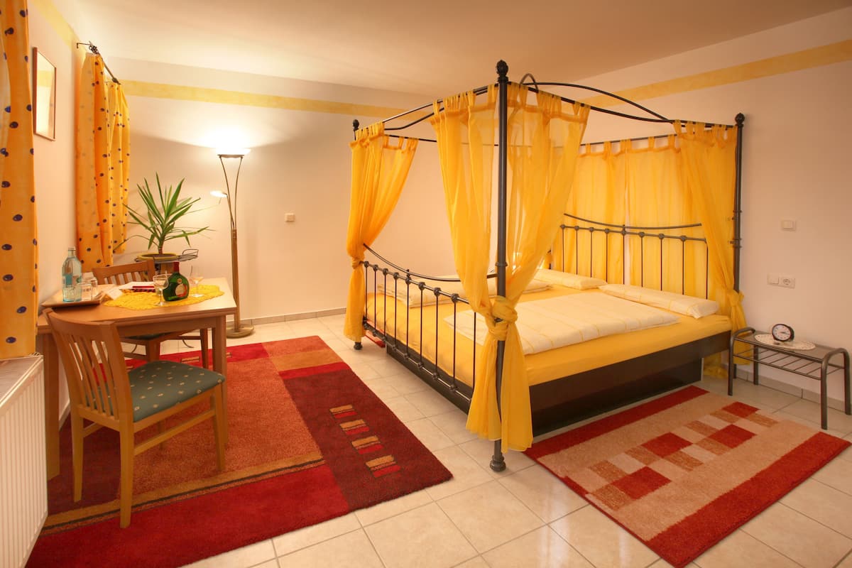 A four-poster bed with yellow curtains, a wooden chair, a small table with a plant, and a red and beige checkered rug.