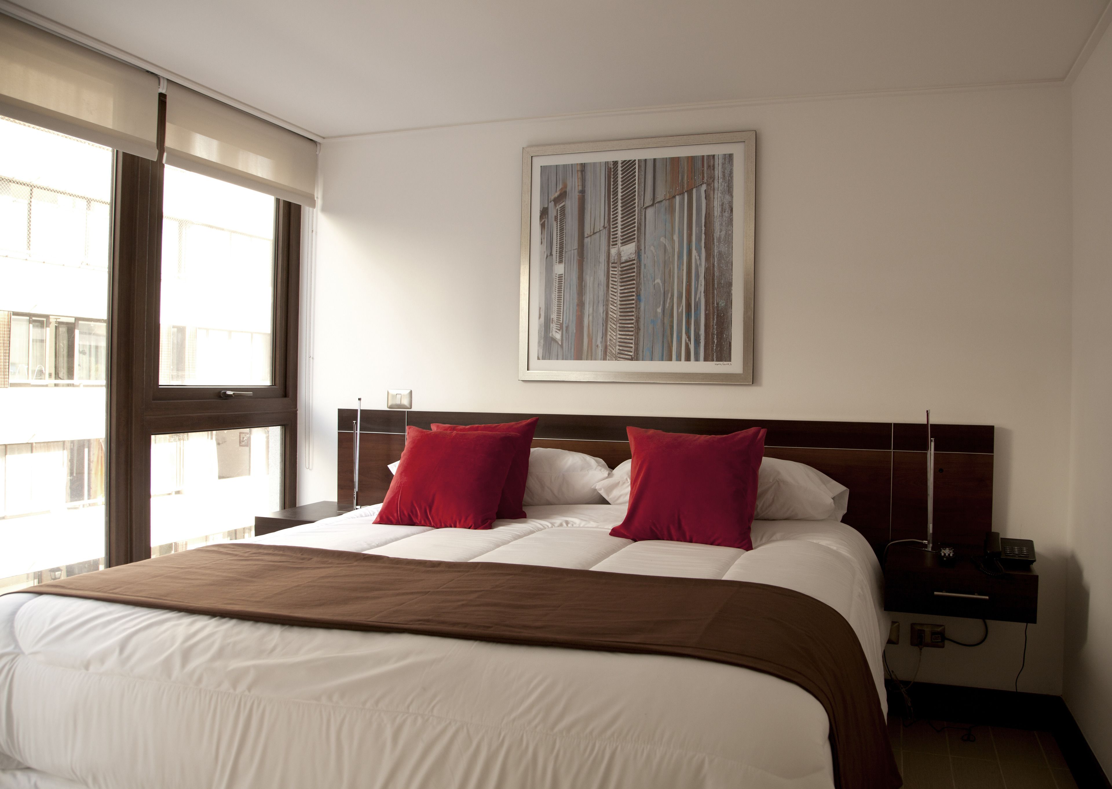 Ein Hotelzimmer mit einem großen Bett, zwei roten Kissen, einer braunen Tagesdecke, einem Nachttisch mit Telefon und einem Gemälde an der Wand.