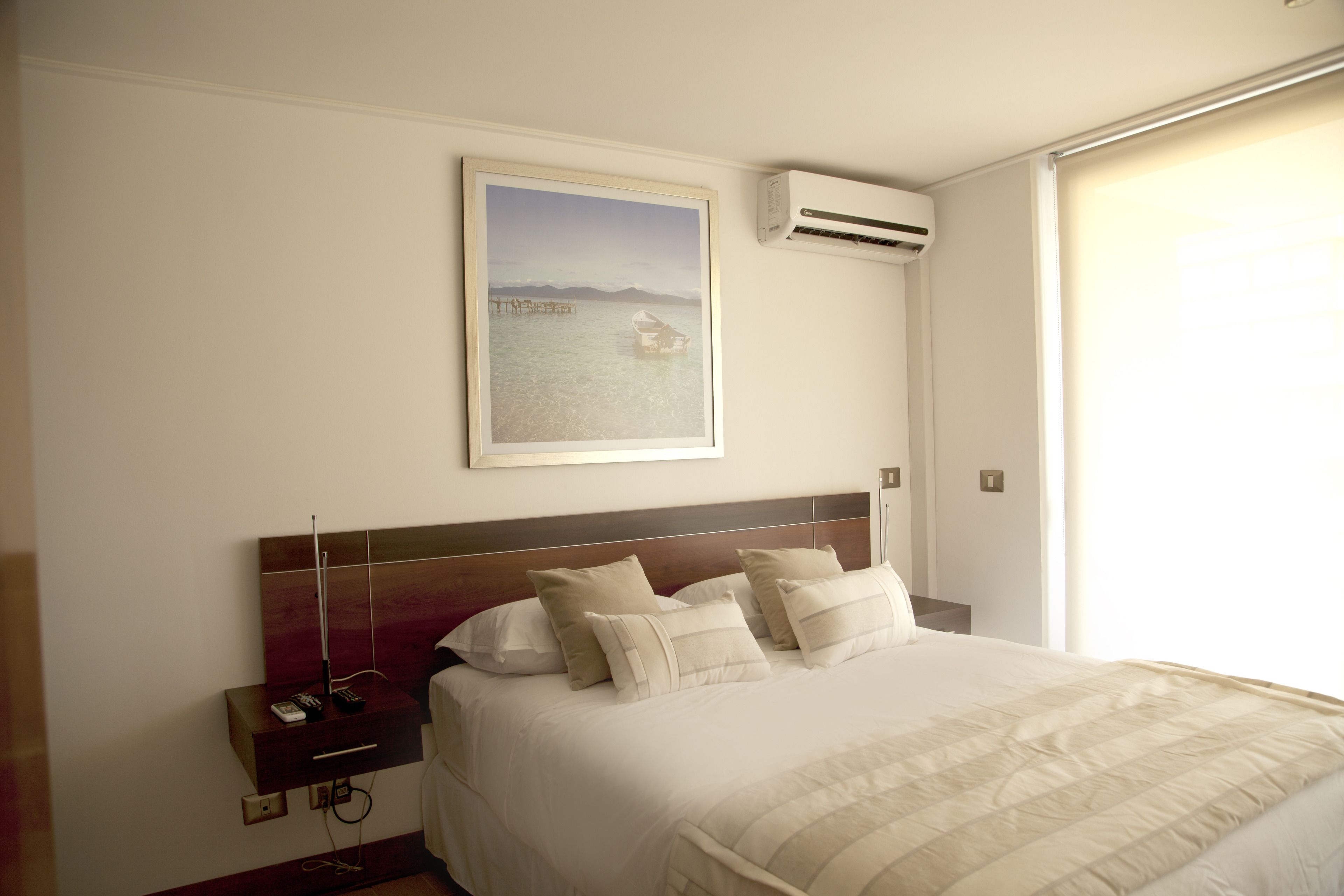 A hotel room with a large bed, a nightstand, a painting of a coastal scene, and an air conditioning unit.