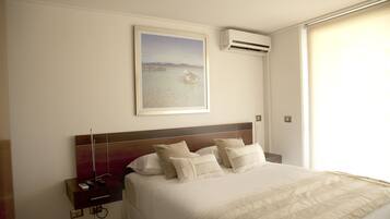 A hotel room with a large bed, a nightstand, a painting of a coastal scene, and an air conditioning unit.