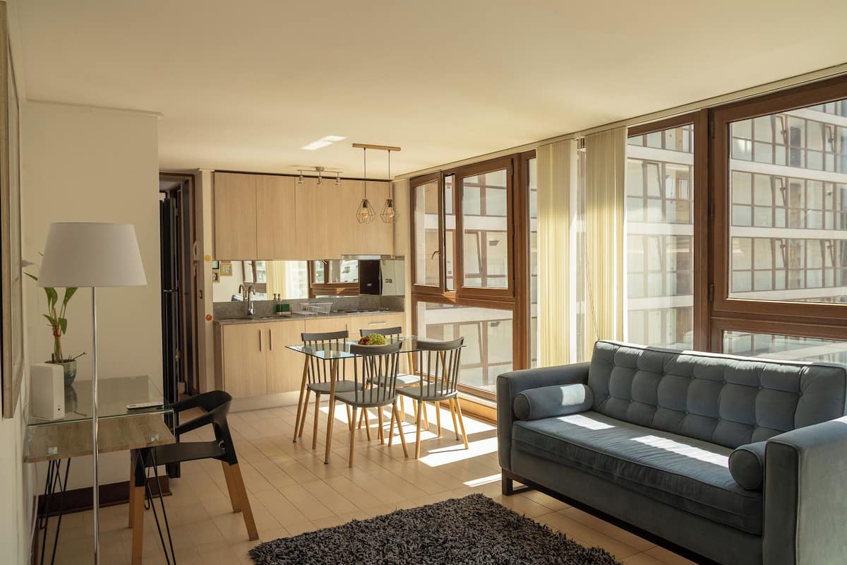 Una sala de estar moderna con un sofá gris, una mesa de comedor de vidrio con sillas, y una cocina al fondo.
