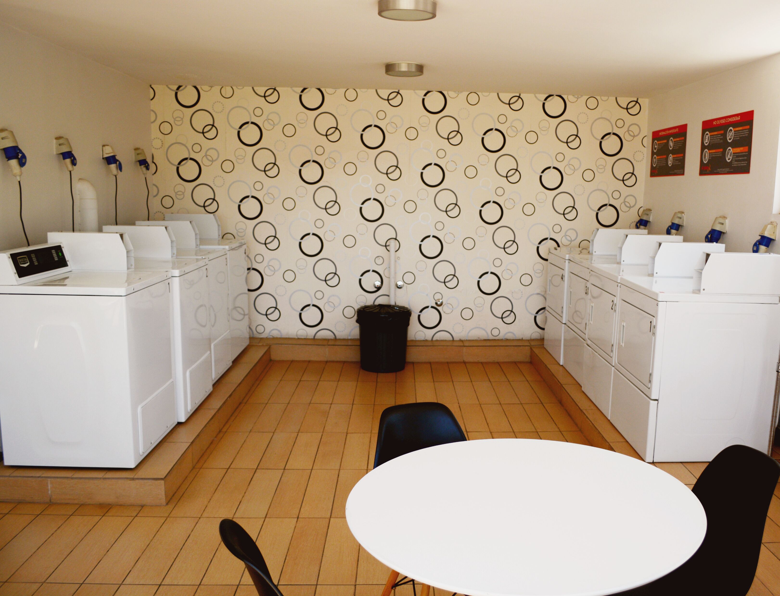 Une buanderie avec un lave-linge blanc, un sèche-linge et une table avec des chaises. On aperçoit un bac à ordures et un mur à motifs à l’arrière-plan.