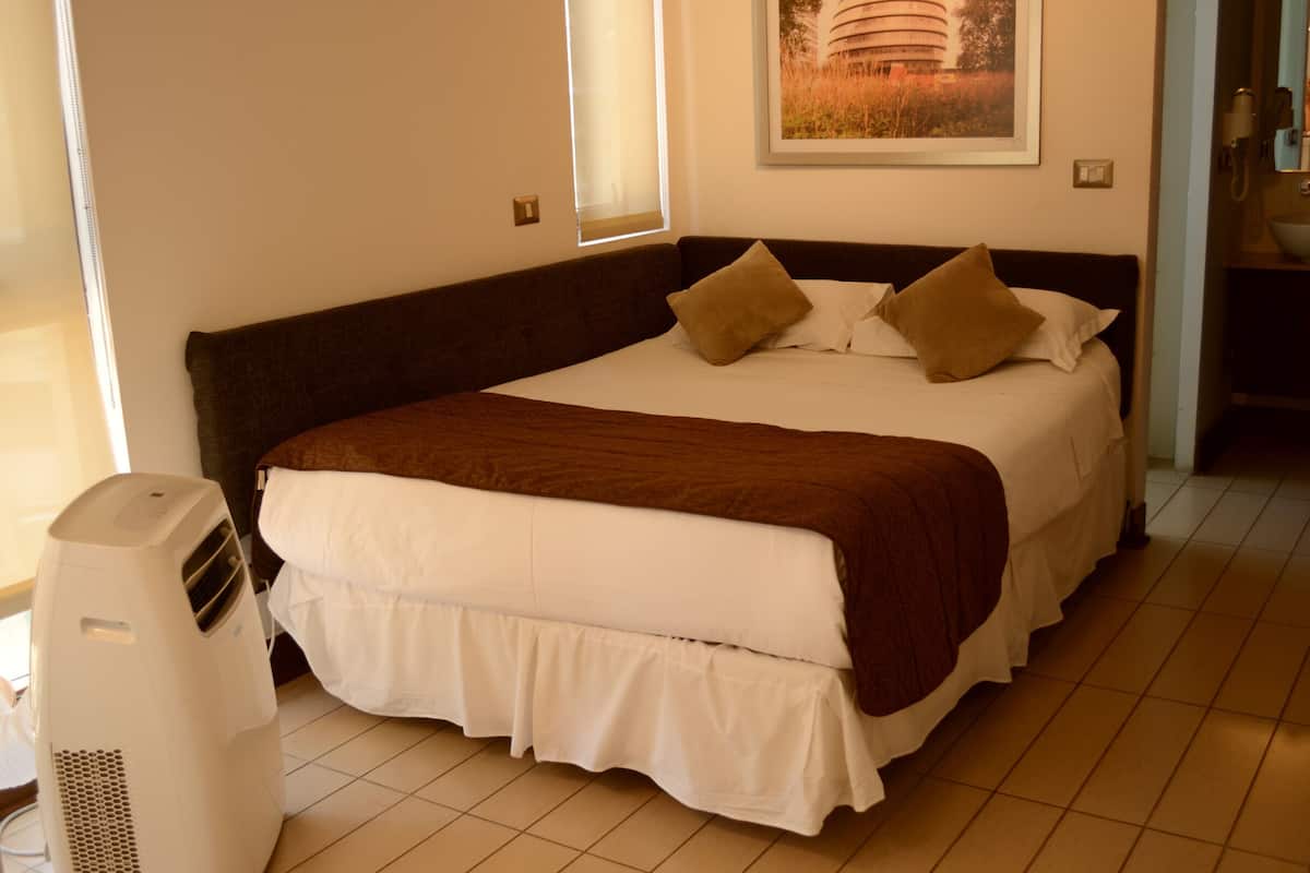 Una cama bien hecha con cabecera marrón, ropa de cama blanca y dos almohadas marrones. Se observa una unidad de aire acondicionado blanca a la izquierda de la cama.