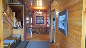 Interior de una cabaña de madera con una escalera, un comedor con una mesa larga y sillas, y una zona de bar con taburetes.