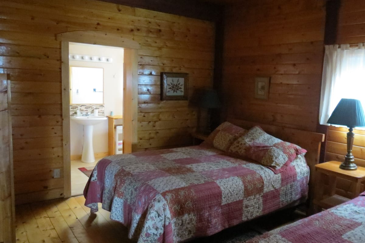 Quarto de cabana de madeira com cama, mesa de cabeceira, luminária e banheiro visível por uma porta aberta.