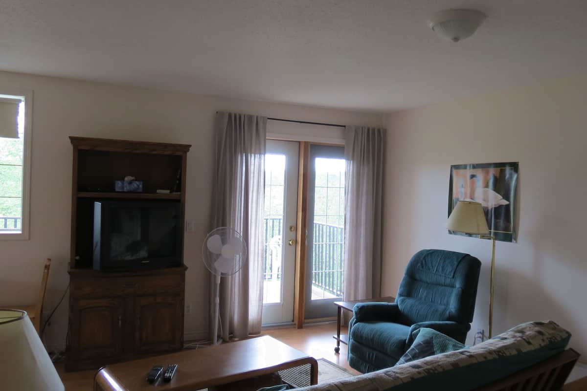 Uma sala de estar com televisão, ventilador, cadeira, mesa e luminária.
