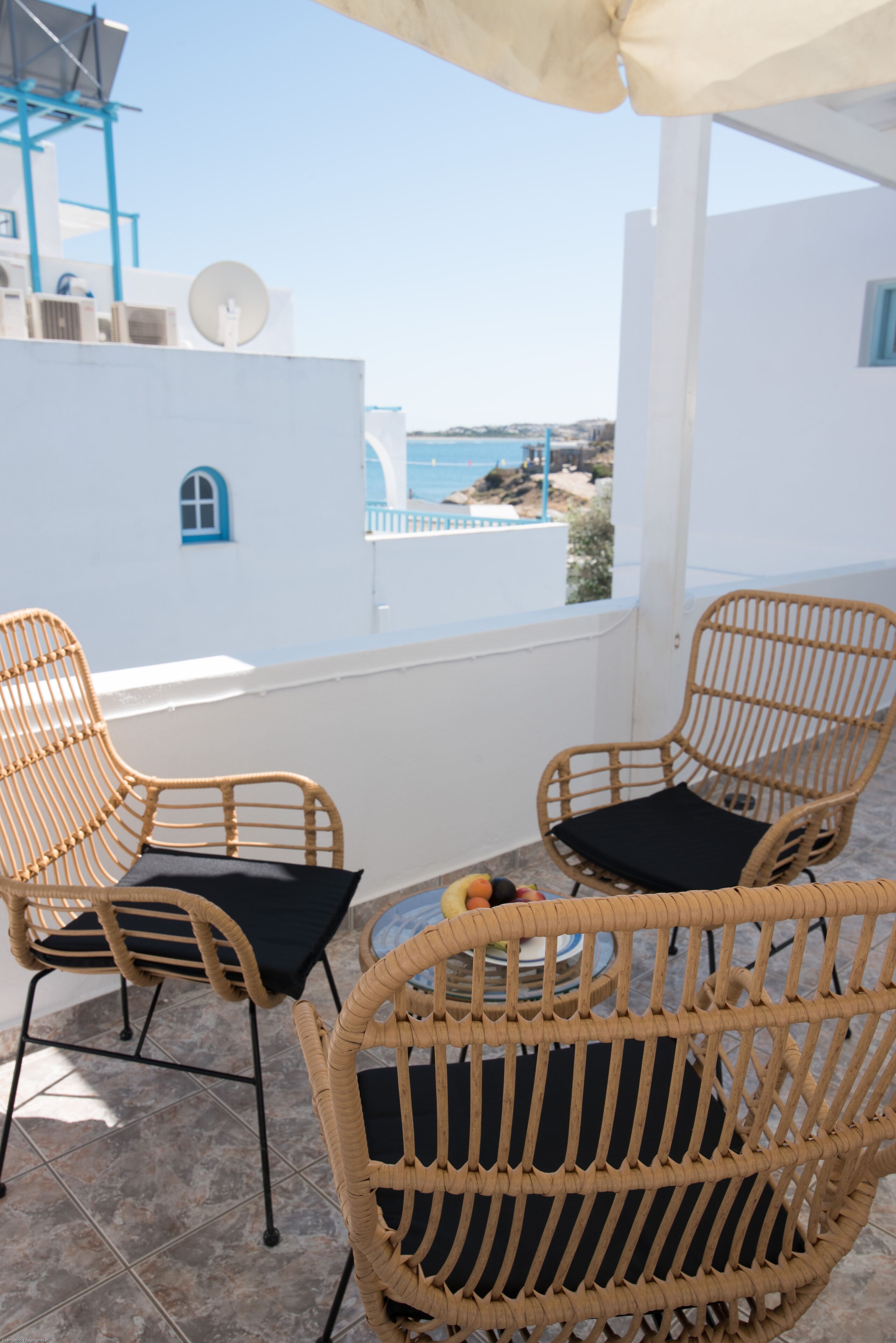 A balcony with wicker chairs and a table, overlooking a coastal view with buildings and a clear sky.