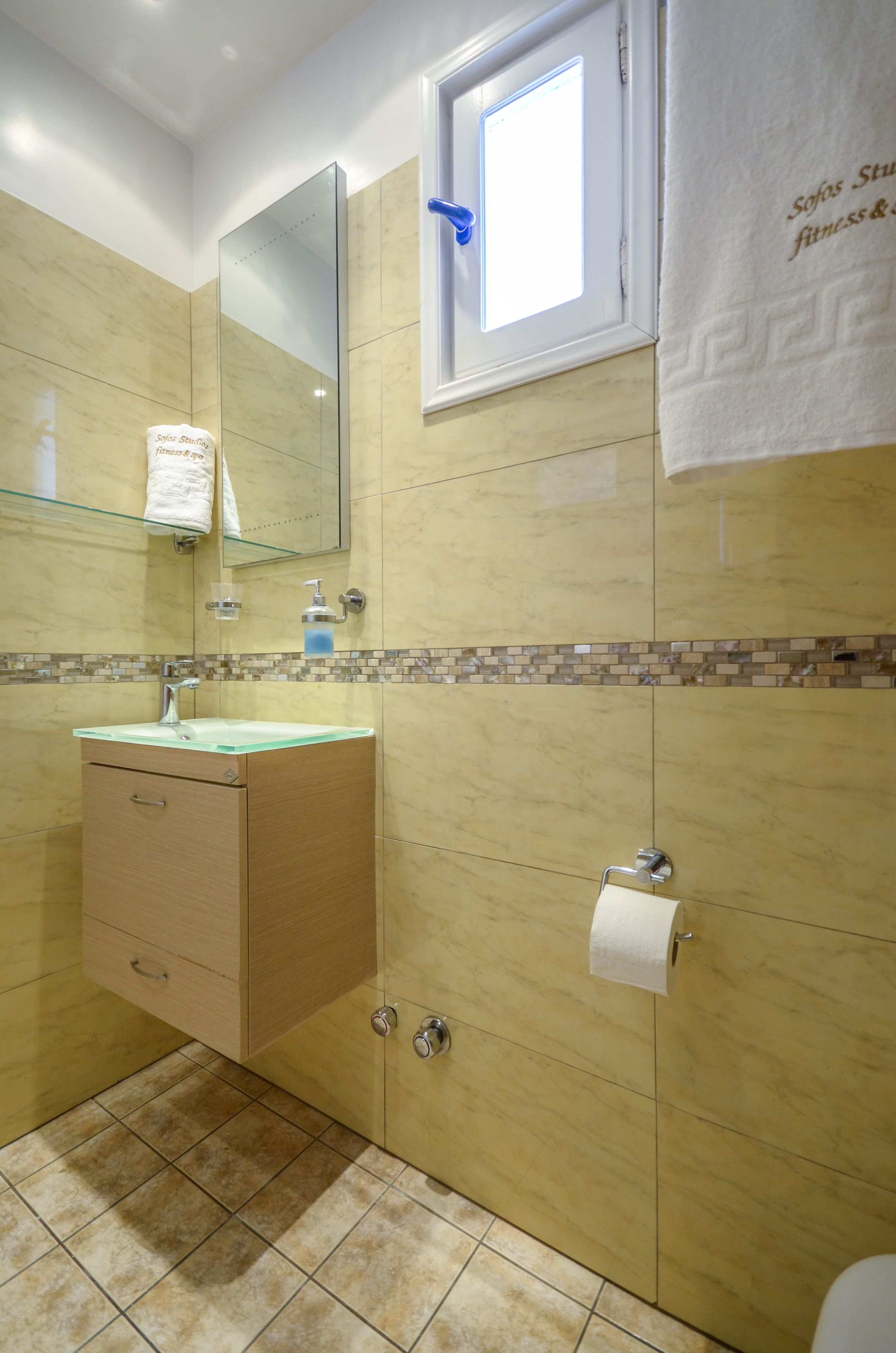 A bathroom with a glass sink, wooden cabinet, mirror, and a window with a towel hanging on the wall.