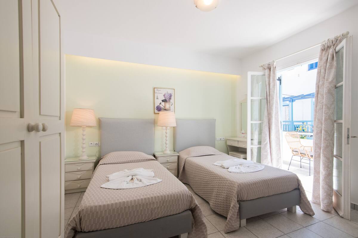 A hotel room with two beds, bedside lamps, a small table, a wall-mounted picture, and a balcony with a chair.