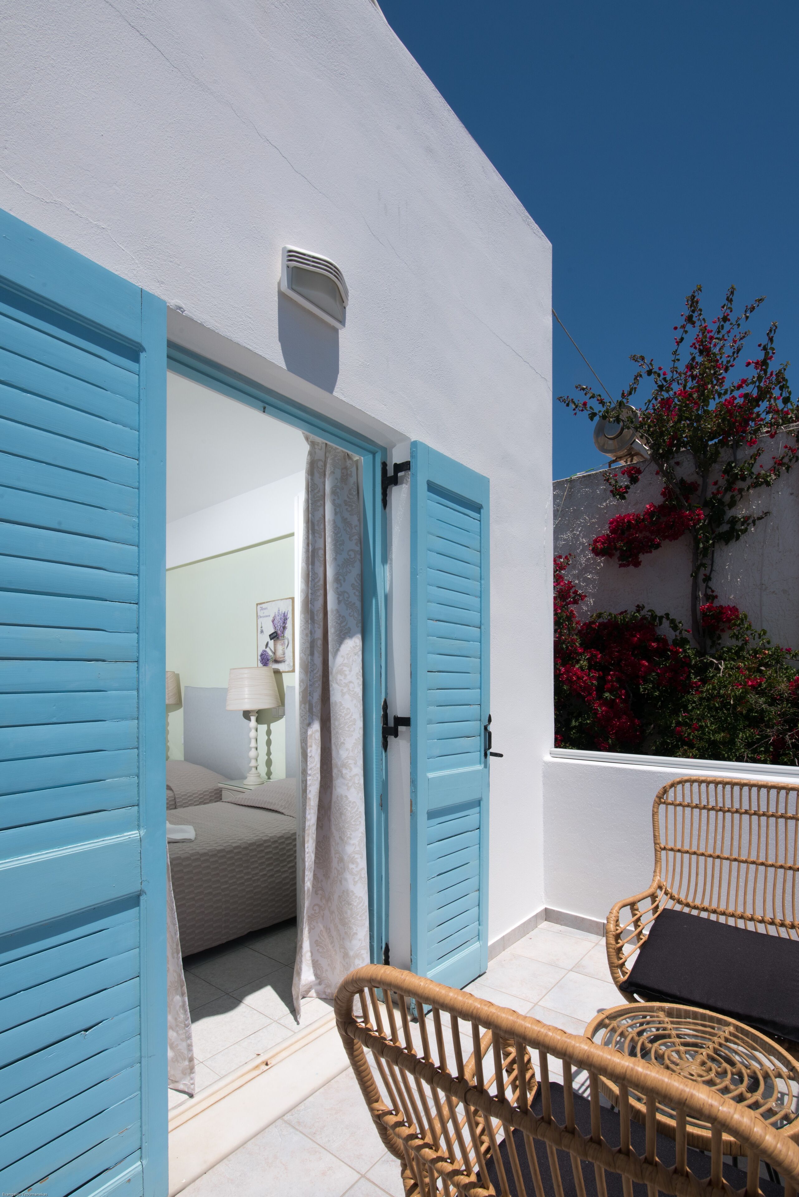 A room with a bed, a window with blue shutters, a white wall, and a view of a tree with red flowers.