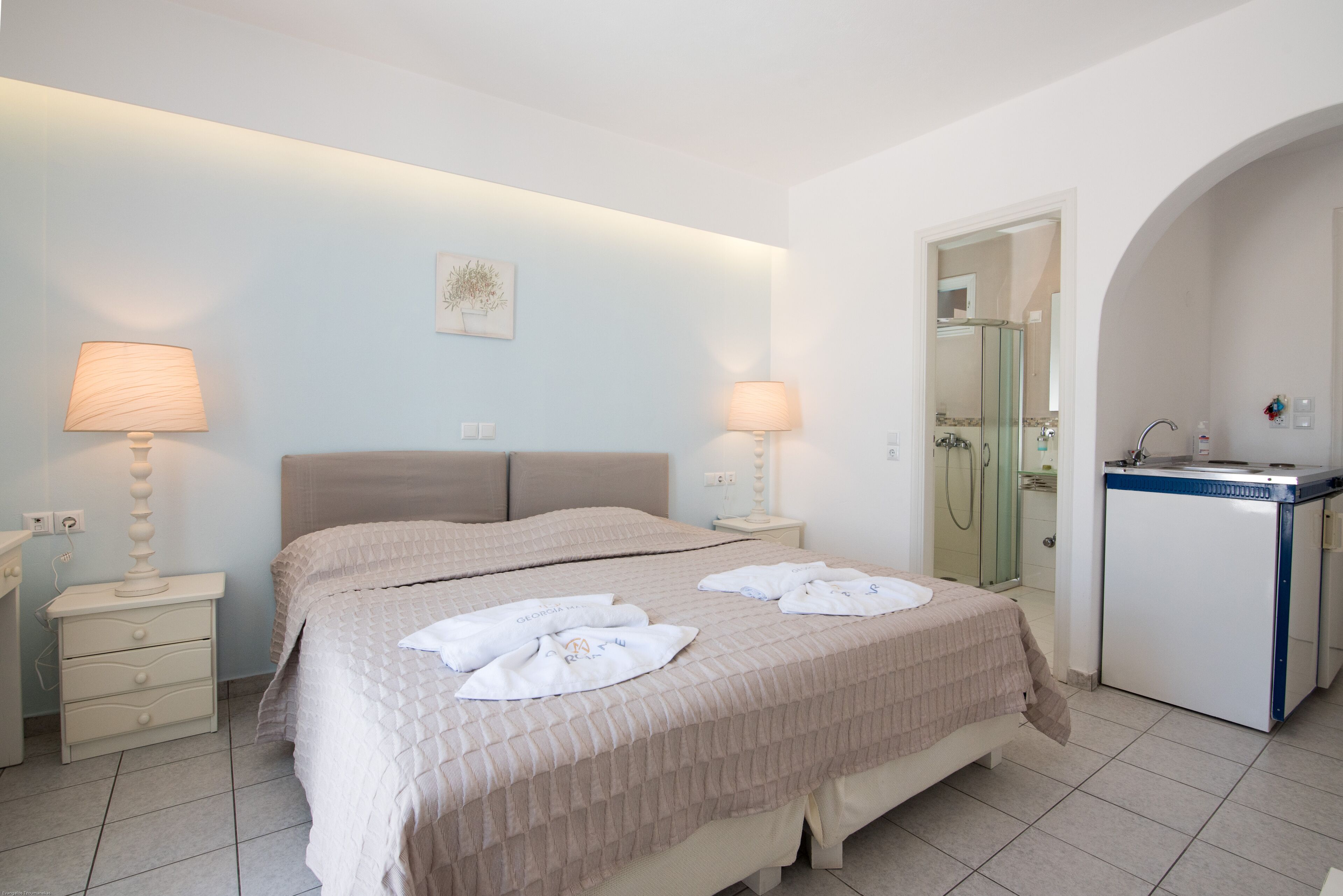 A hotel room with a bed, two bedside lamps, a small desk with a mirror, and a bathroom visible through an archway.