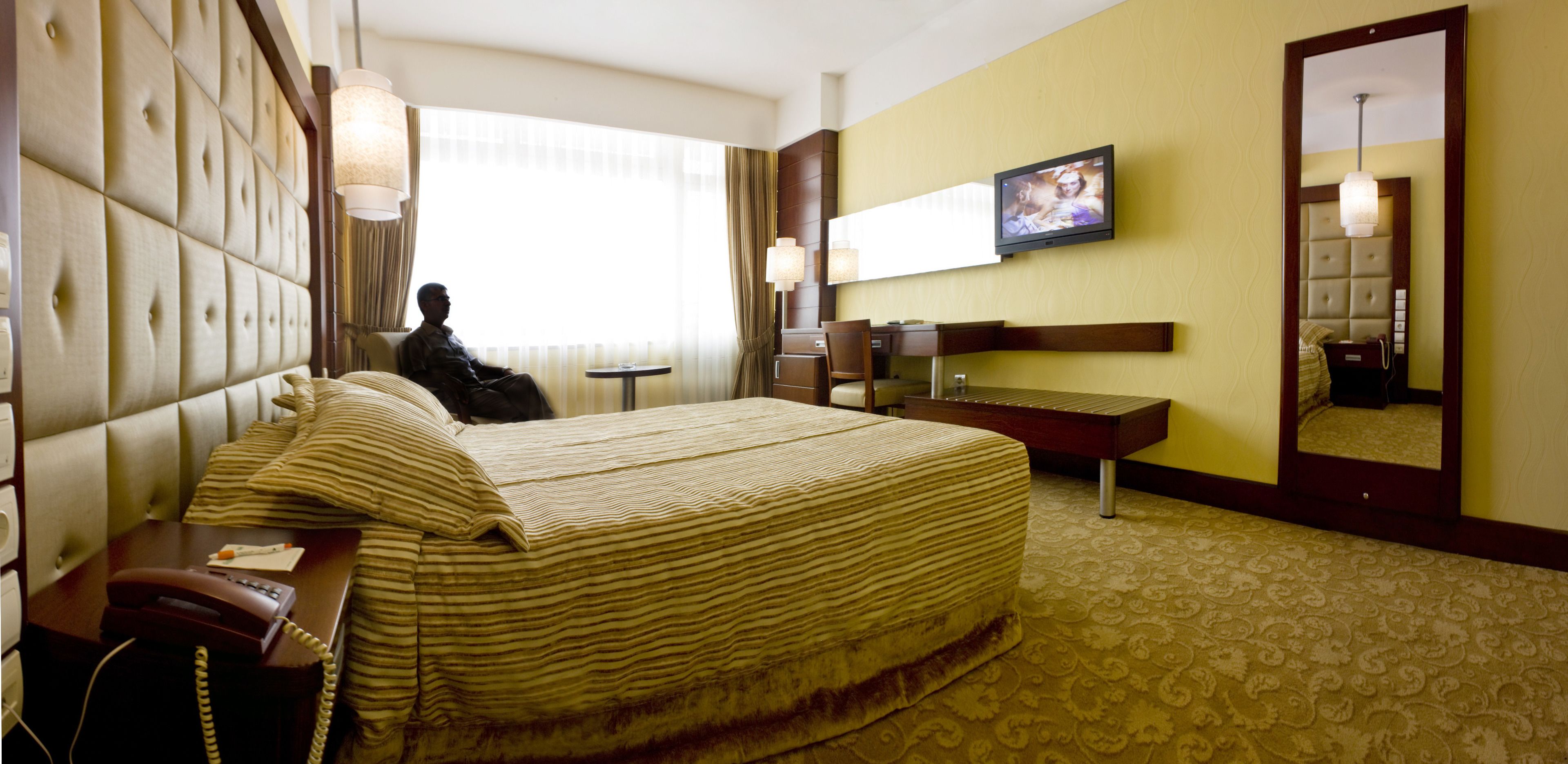 Una habitación de hotel con una cama grande, una mesita de noche con teléfono, un escritorio con silla, un televisor montado en la pared y un espejo amplio.