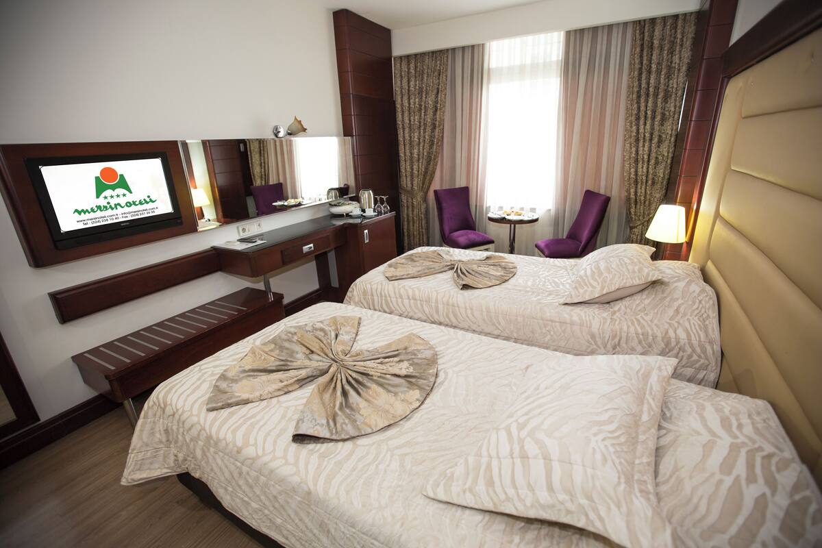 Quarto de hotel com duas camas, uma escrivaninha, uma televisão, uma cadeira e uma mesa pequena.