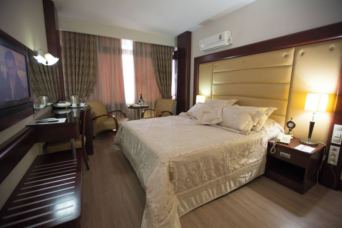 Quarto de hotel com uma cama grande, uma escrivaninha com computador, uma cadeira e uma televisão.