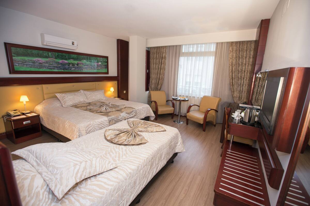 Quarto de hotel com duas camas, uma escrivaninha, uma cadeira, televisão e uma janela com cortinas.