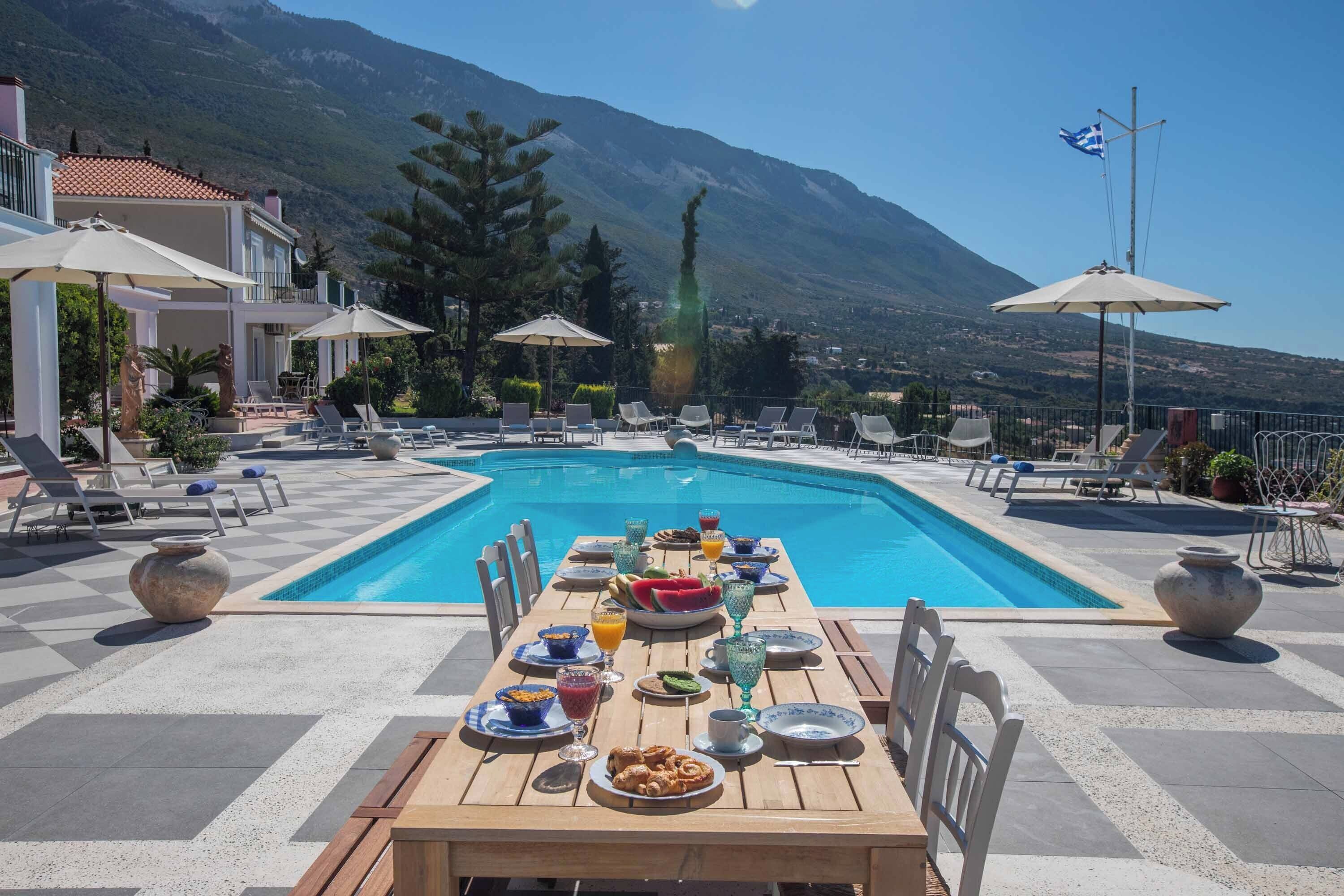 A poolside dining area with a table set for breakfast, overlooking a mountain and a pool.
